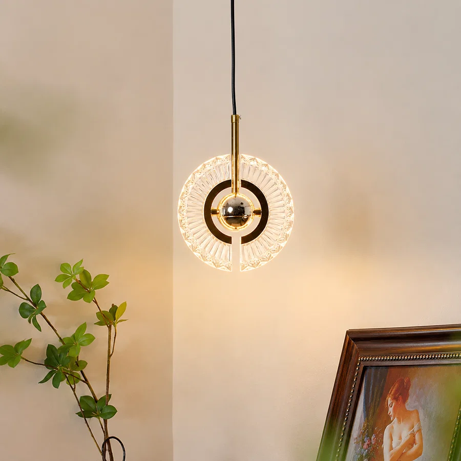 A modern circular pendant light with a gold-finished central rod and clear faceted glass rings, suspended against a warm beige wall. Part of a green plant and a framed picture are visible in the room setting.