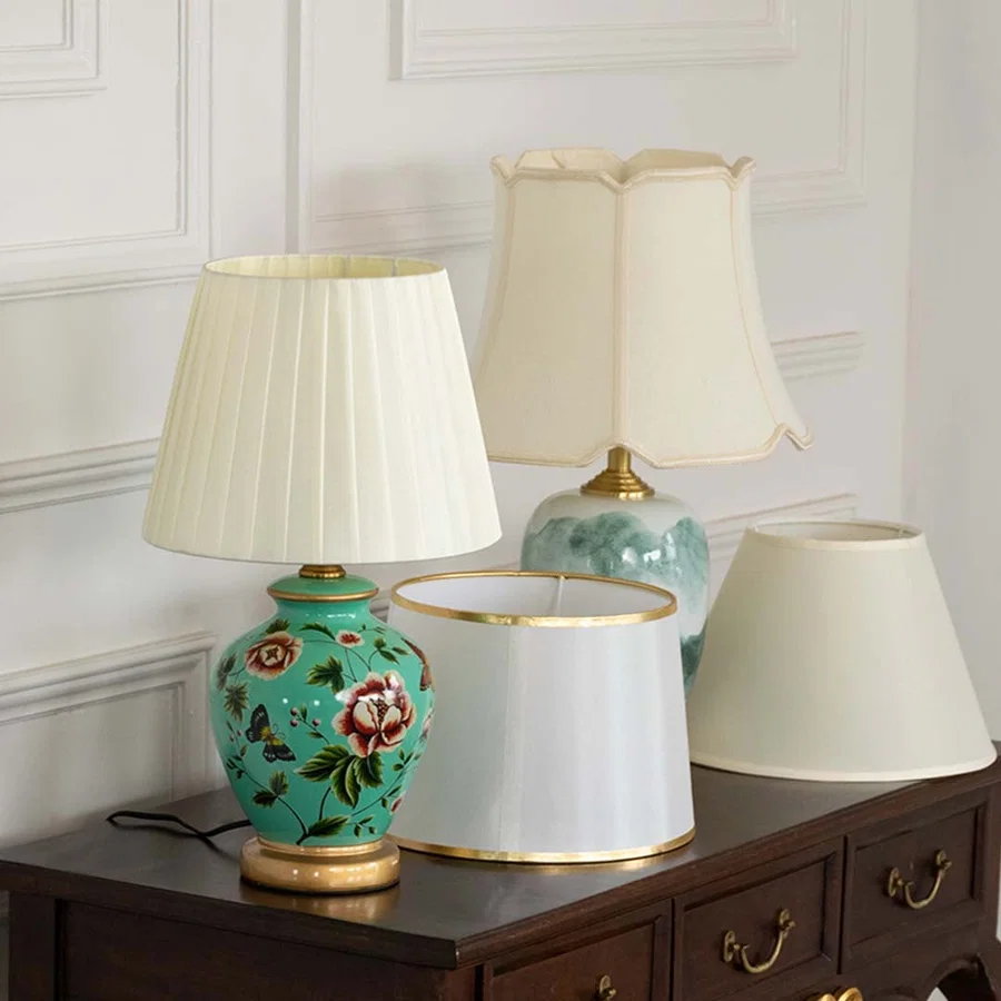 Green floral ceramic table lamp with pleated shade displayed on wooden console beside pastel lampshades