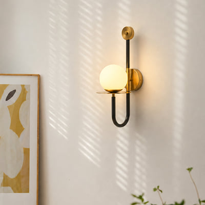 A modern wall sconce with a glowing frosted glass globe and matte black U-shaped arm, mounted on a white wall with soft shadow patterns from nearby leaves. A framed abstract print is partially visible on the left.