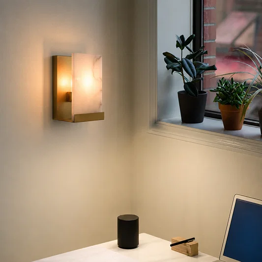 A modern wall-mounted sconce with a rectangular gold-toned metal frame and a glowing warm white marble-look glass shade, installed on a beige wall in a room setting next to a window with potted plants.