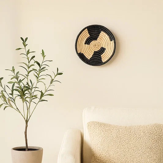 A handwoven natural and black rattan wall decor plate styled above a marble table with a tall black vase and geometric sculpture creating warm modern home decor vibes.