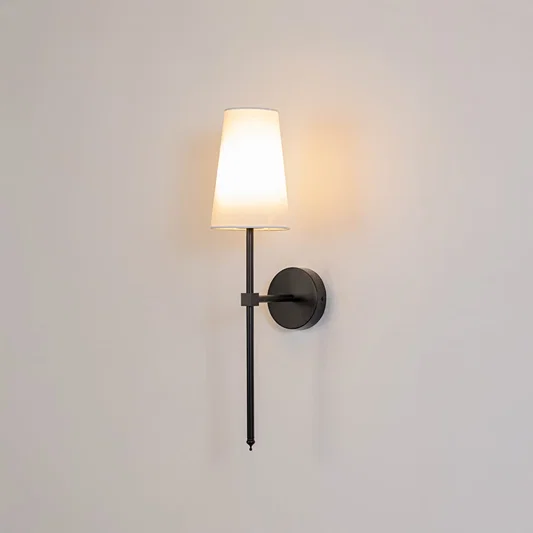 Illuminated white fabric drum shade wall light with tall black metal rod emitting warm ambient glow on white background.