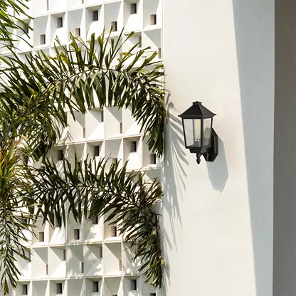 Black metal outdoor wall lantern with clear textured glass mounted on a white exterior wall with greenery adding refined architectural outdoor styling.