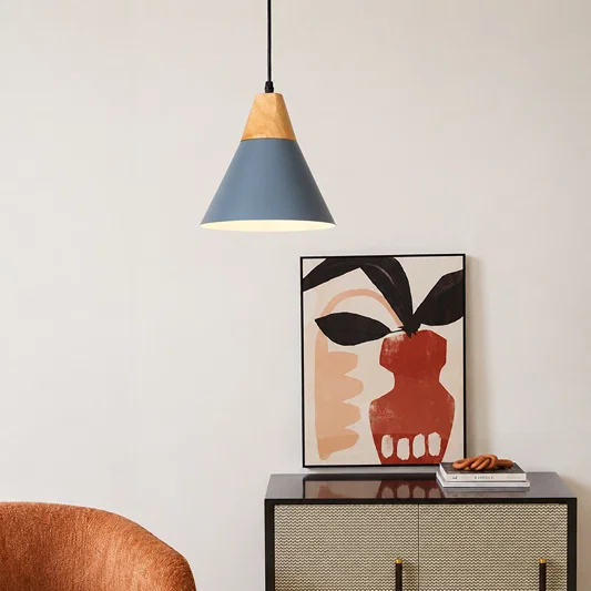 A modern pendant light with a blue-gray and natural wood shade hangs above a black cabinet in a styled interior. A framed abstract art print and a small stack of books sit on the cabinet against a light neutral wall.