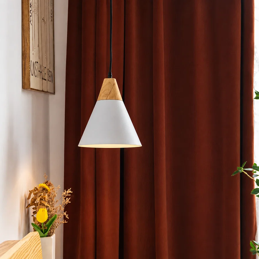 A modern pendant light with a white cone shade and natural wood accents hangs in front of dark rust-brown curtains in a styled interior setting.