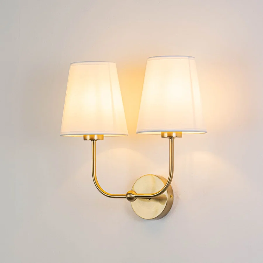 Gold metal double wall sconce with white fabric shades illuminated on a bright white background for clarity.
