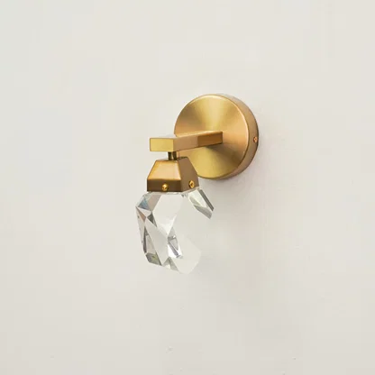 A modern wall-mounted light fixture with a brushed gold circular backplate, a geometric gold metal arm, and a faceted clear crystal shade, shown against a plain off-white wall.