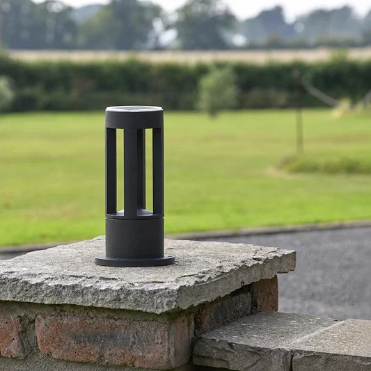 A modern matte black cylindrical bollard light with vertical slotted cutouts, mounted on a stone pillar in an outdoor garden setting with a blurred green lawn and trees in the background.