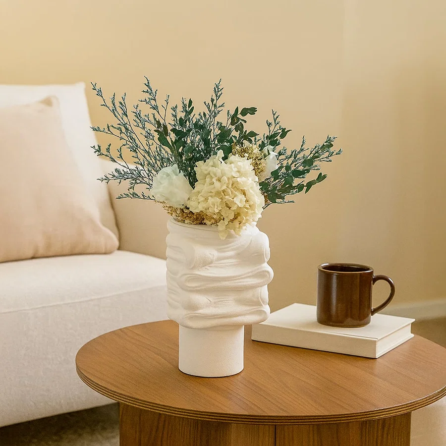 White sculptural ceramic vase with modern textured design styled on a wooden coffee table with flowers beside a sofa in a cozy neutral living room.