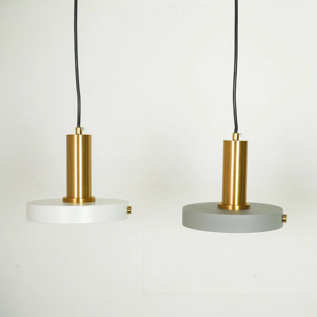 Two modern pendant lights with cylindrical gold brass centers and circular shades, one in white and one in gray, hanging against a plain white wall.