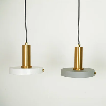 Two modern pendant lights with cylindrical gold brass centers and circular shades, one in white and one in gray, hanging against a plain white wall.