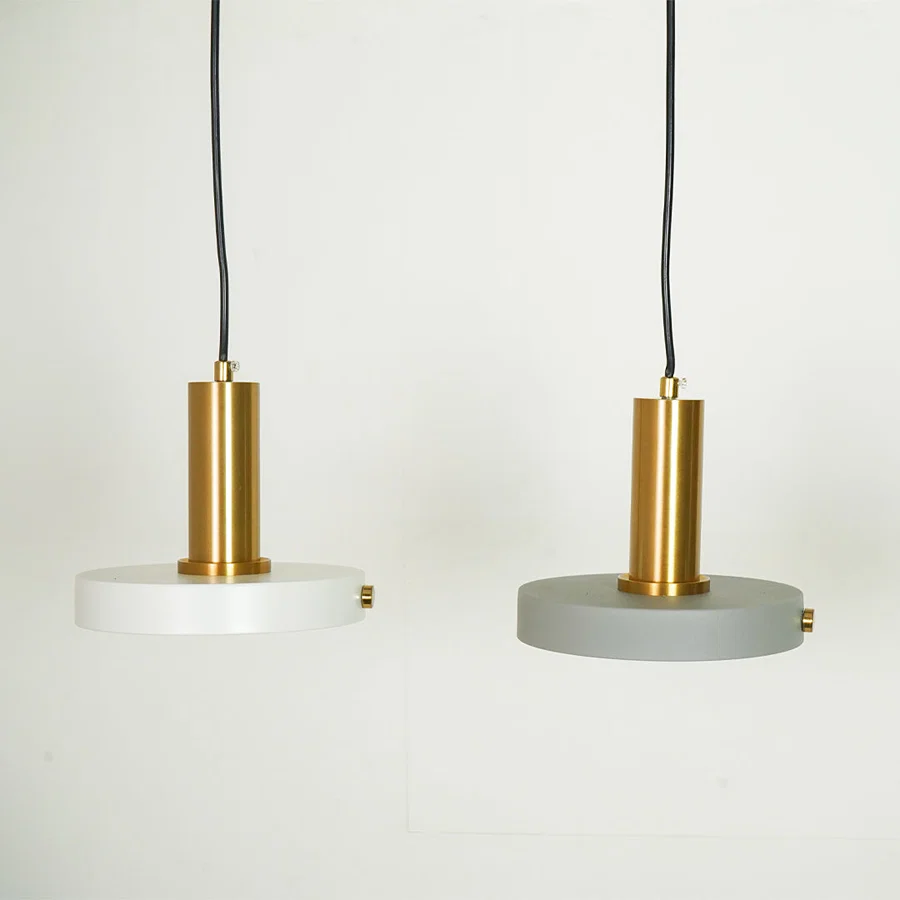 Two modern pendant lights with cylindrical gold brass centers and circular shades, one in white and one in gray, hanging against a plain white wall.