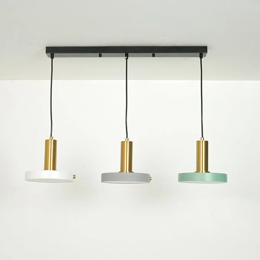 Three modern pendant lights hanging from a black ceiling bar. Each light has a cylindrical gold-toned metal shade, a black cord, and a round shade at the bottom in white, light gray, and mint green.