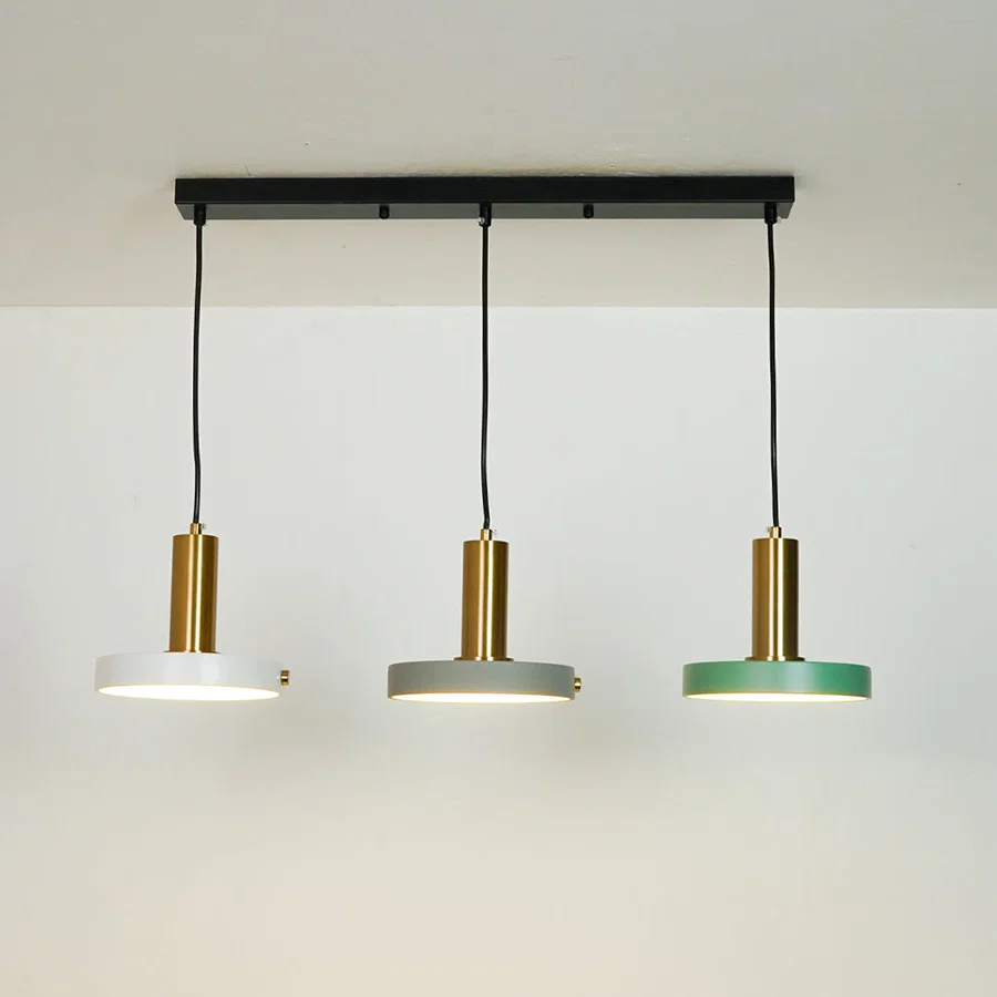 Three-light linear ceiling pendant fixture with a black horizontal bar, black cords, and three round shades in white, gray, and green with brushed gold centers, shown against a plain off-white wall.