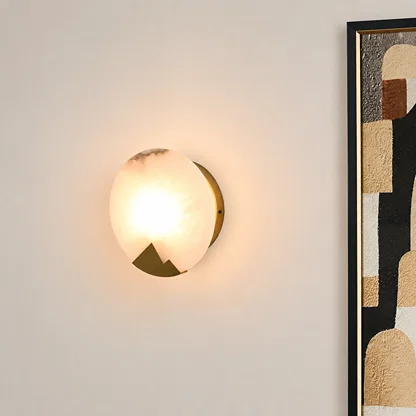 A modern wall sconce light fixture with a glowing frosted glass globe and a gold-toned geometric metal base, mounted on a light beige wall next to a framed abstract art print.