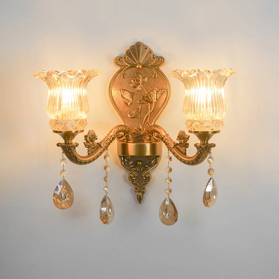 An ornate double-arm wall sconce with a gold-finished metal frame, featuring two fluted clear glass shades, hanging teardrop crystal drops, and a decorative crest plate on the backplate, shown illuminated against a plain light gray wall.