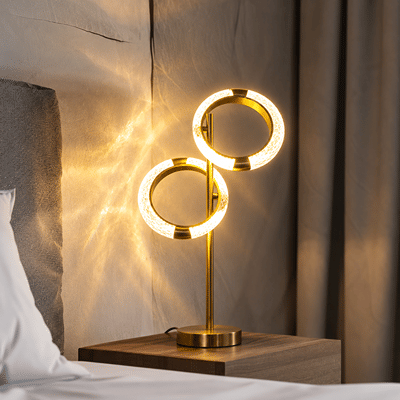 A modern table lamp with two glowing circular rings on a slim gold stand, placed on a wooden bedside table next to a bed in a softly lit bedroom.