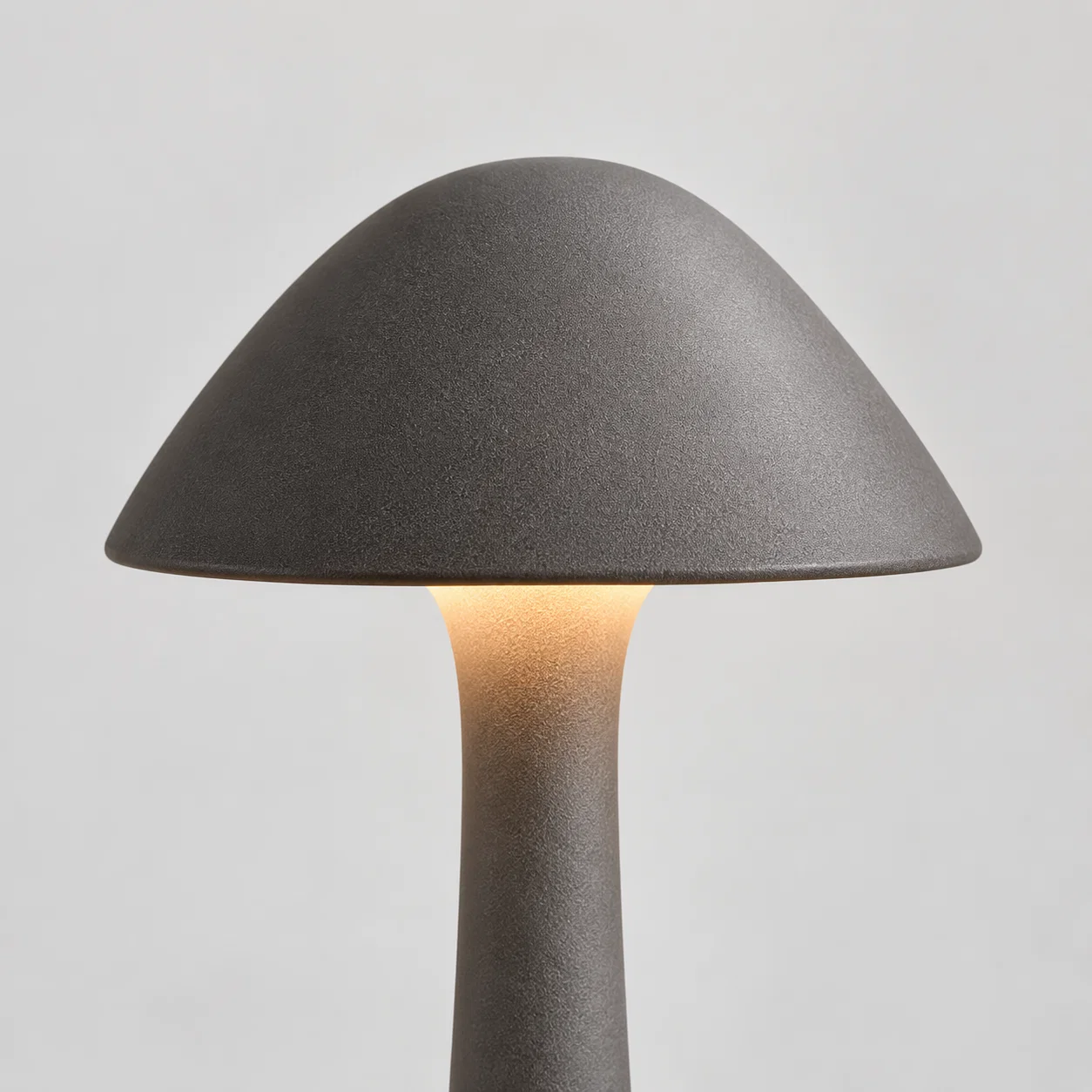 A mushroom-shaped table lamp with a textured dark gray ceramic shade and stem, glowing with a warm light from the center, set against a neutral light gray background.