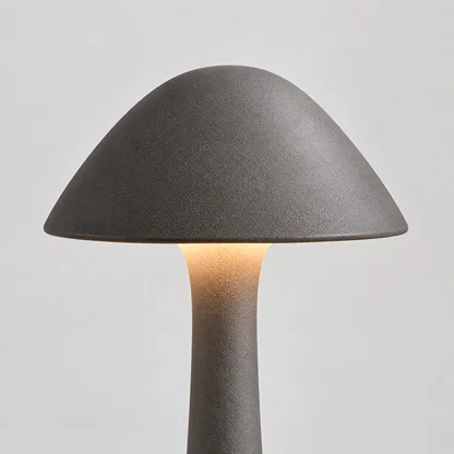A mushroom-shaped table lamp with a textured dark gray ceramic shade and stem, glowing with a warm light from the center, set against a neutral light gray background.