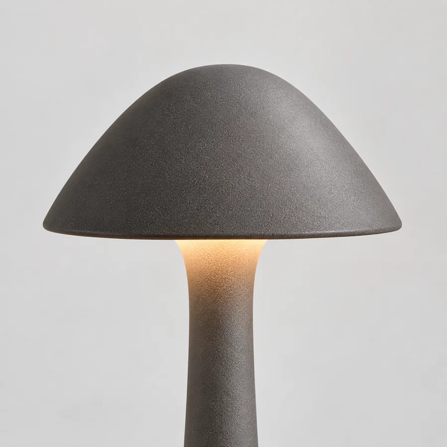 A mushroom-shaped table lamp with a textured dark gray ceramic shade and stem, glowing with a warm light from the center, set against a neutral light gray background.
