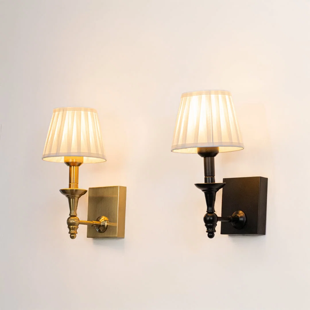 Black wall sconce with white pleated shade illuminated on a clean white background showing warm soft glow.