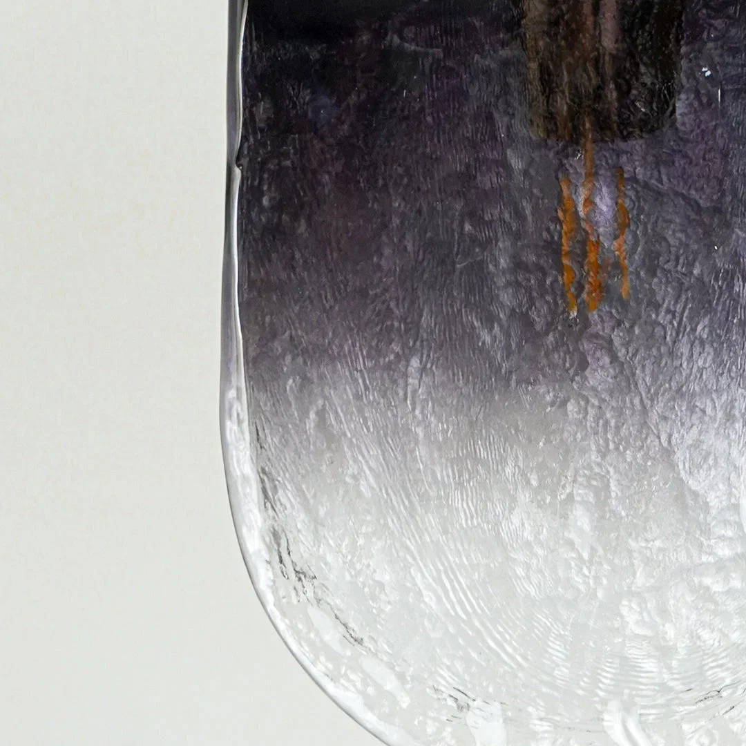 Textured glass pendant light with dark to clear gradient finish and gold bulb holder placed against neutral wall background closeup showcasing elegant lighting design.
