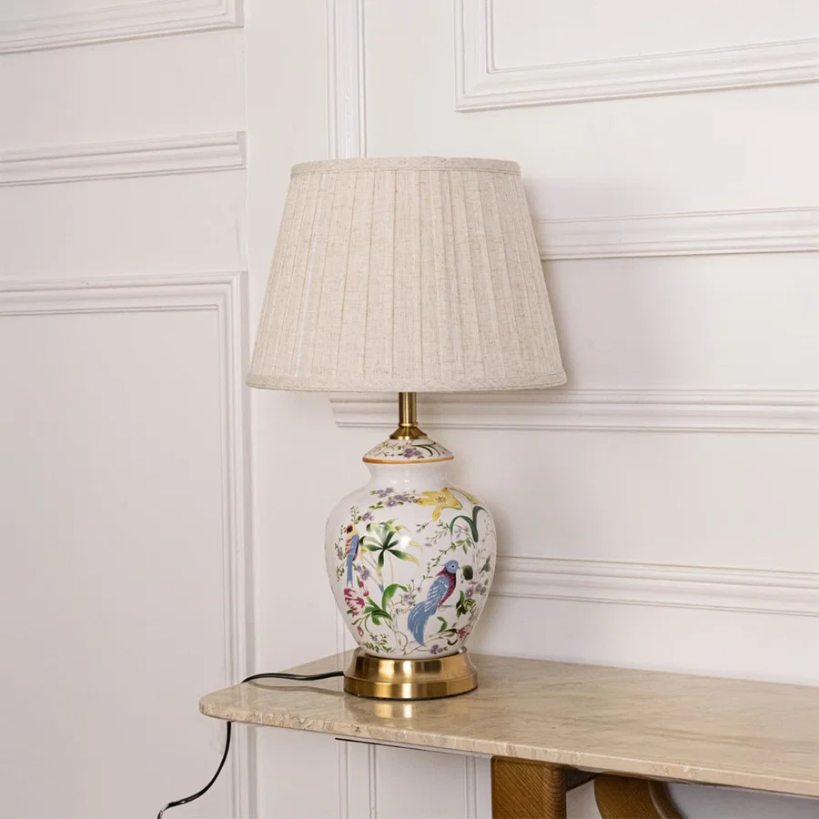 Decorative ceramic table lamp with colorful hand painted floral design gold base and pleated fabric shade on off white background.
