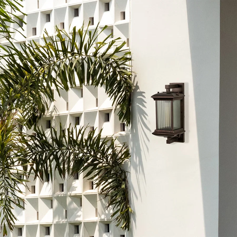 A stylish bronze outdoor lantern fixed on a white architectural wall glowing softly and adding character and charm to the surrounding outdoor environment.
