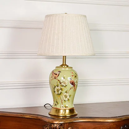 Decorative green ceramic table lamp with bird and floral design brass base and fabric shade on soft off white background.