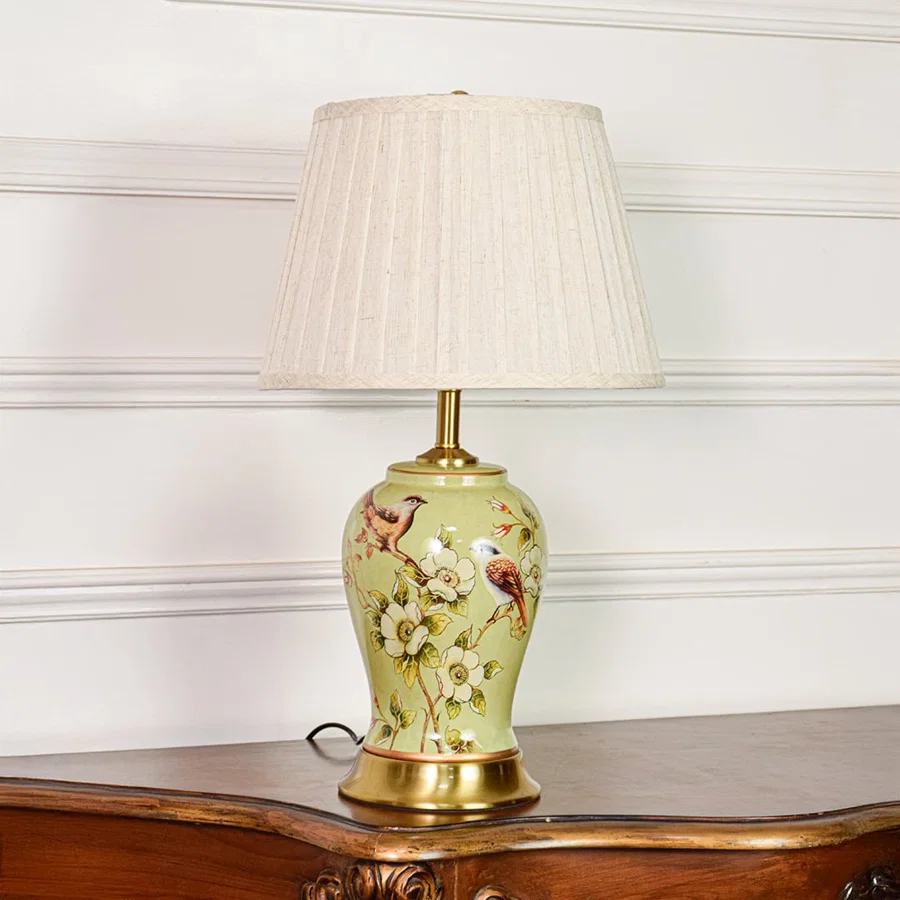 Decorative green ceramic table lamp with bird and floral design brass base and fabric shade on soft off white background.