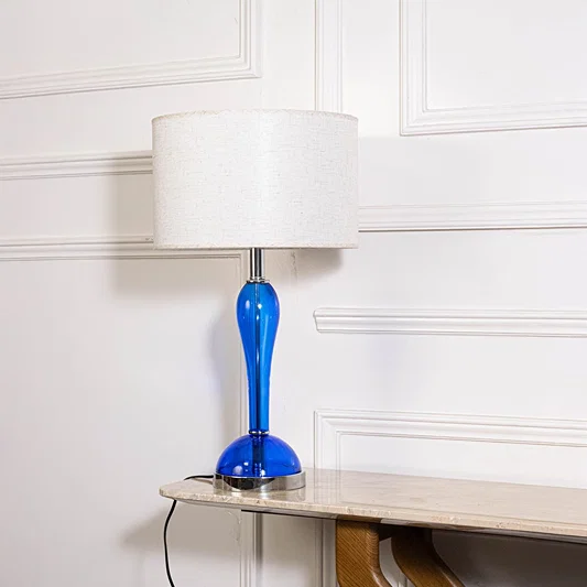 Blue glass lamp with lights off highlighting glossy glass body and fabric shade on plain white background.
