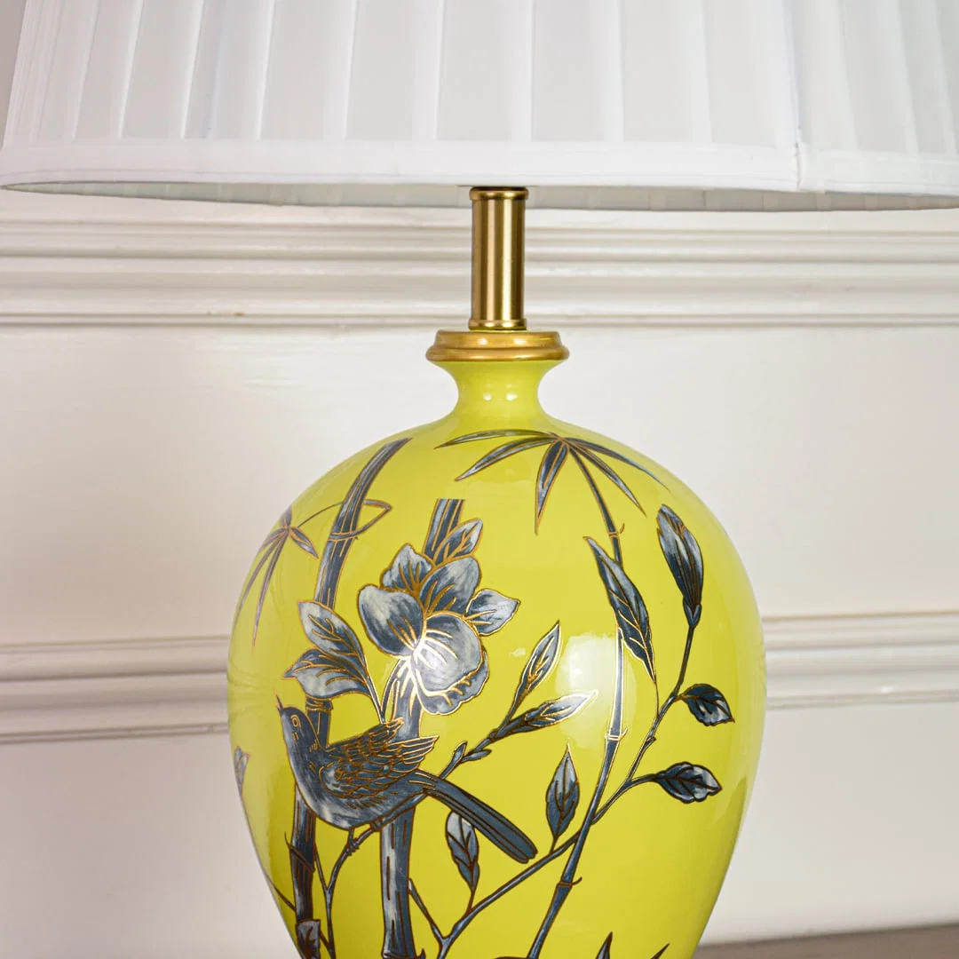Closeup of yellow ceramic table lamp highlighting hand painted floral bird details smooth glaze finish and gold metal base.