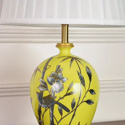 Closeup of yellow ceramic table lamp highlighting hand painted floral bird details smooth glaze finish and gold metal base.