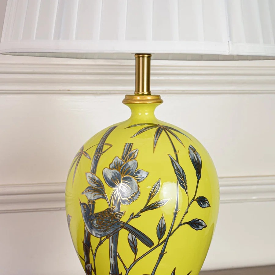 Closeup of yellow ceramic table lamp highlighting hand painted floral bird details smooth glaze finish and gold metal base.