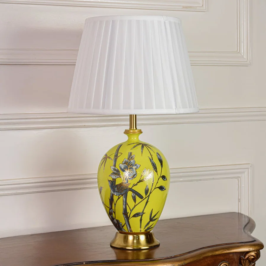 Decorative yellow ceramic table lamp with floral bird artwork gold metal base and pleated fabric shade on off white background.
