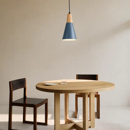 A modern dining setup featuring a round light oak table, two dark wood chairs with black seats, and a blue and wood conical pendant light hanging above against a neutral beige wall.