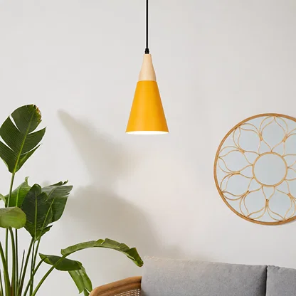 A modern cone-shaped pendant light with a yellow body and a light wood top, hanging in a minimalist room setting next to a green plant and a gray sofa.