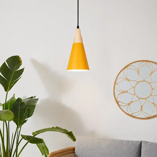 A modern cone-shaped pendant light with a yellow body and a light wood top, hanging in a minimalist room setting next to a green plant and a gray sofa.