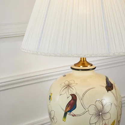 Closeup of ceramic lamp base featuring colorful bird and floral artwork on ivory finish with golden base detailing.