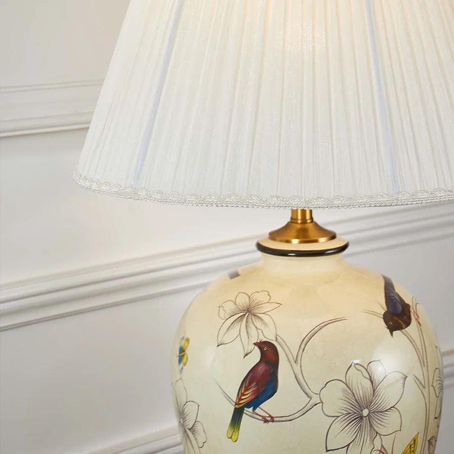 Closeup of ceramic lamp base featuring colorful bird and floral artwork on ivory finish with golden base detailing.