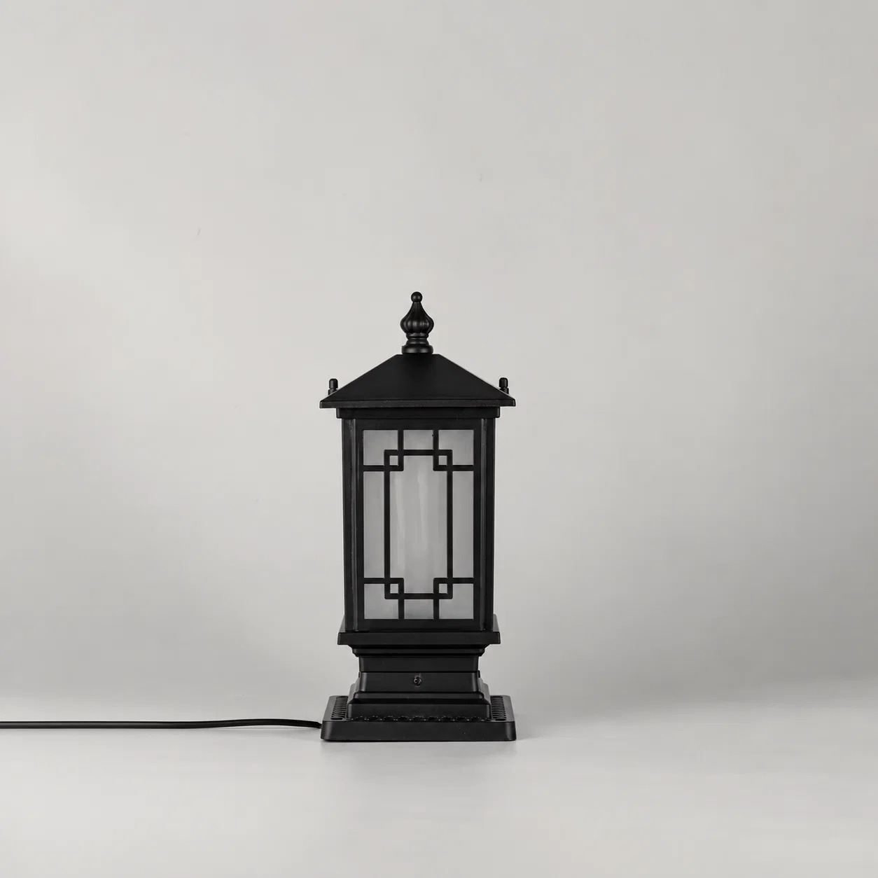 A black outdoor lantern-style table lamp with a square base and geometric glass panels, shown on a light gray studio background with its power cord extending to the left.
