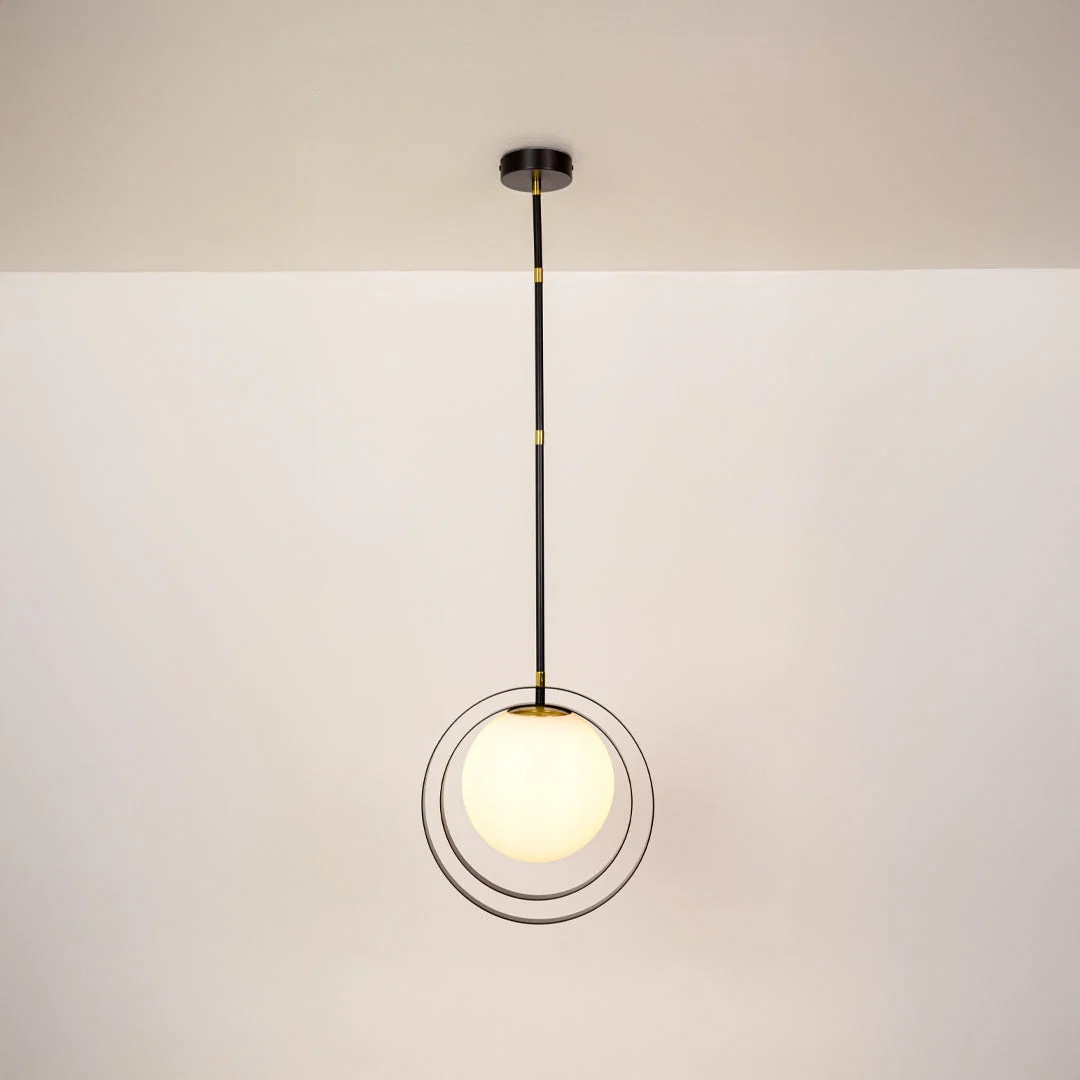A modern pendant light with a black metal rod, gold accents, and a glowing white spherical glass shade suspended in front of two thin black rings, shown against a plain off-white wall and ceiling.