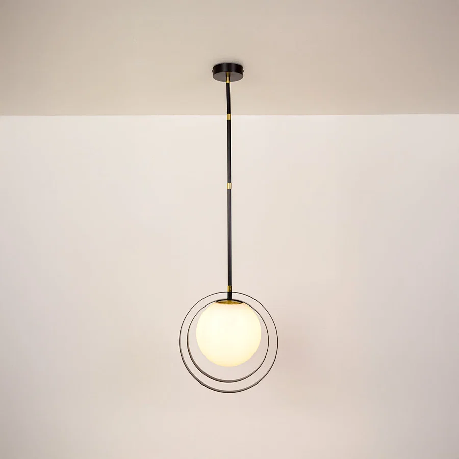 A modern pendant light with a black metal rod, gold accents, and a glowing white spherical glass shade suspended in front of two thin black rings, shown against a plain off-white wall and ceiling.