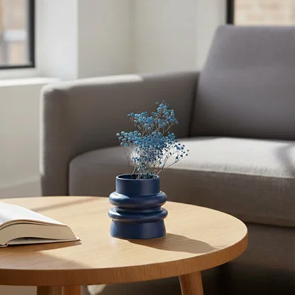 Lifestyle image of a dark blue ribbed ceramic vase styled with blue baby's breath flora on a sunny round coffee table.