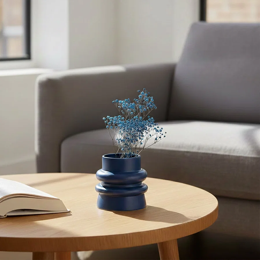 Lifestyle image of a dark blue ribbed ceramic vase styled with blue baby's breath flora on a sunny round coffee table.