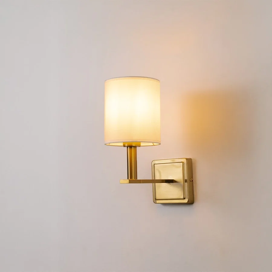 Modern gold wall sconce with white fabric drum shade glowing on white background creating soft warm ambient lighting decor.